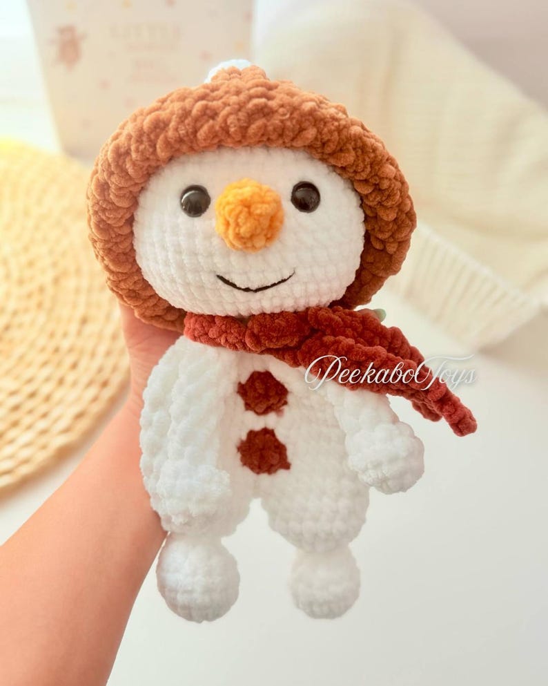 May include: A white crocheted snowman with a brown crocheted hat and a red scarf. The snowman has a carrot nose and black eyes. The snowman is holding a small brown crocheted heart.