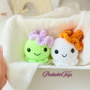 May include: Two crocheted plush toys, one green and one white, each with a smiling face and a flower-like detail on top. The toys are nestled in a white basket with a soft, textured blanket. The text "PeekaboToys" is visible.