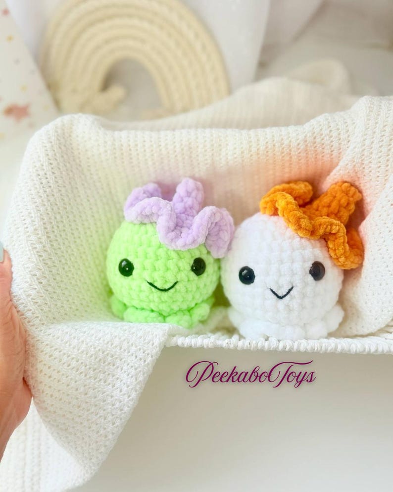 May include: Two crocheted plush toys, one green and one white, each with a smiling face and a flower-like detail on top. The toys are nestled in a white basket with a soft, textured blanket. The text "PeekaboToys" is visible.