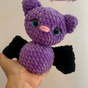 May include: A handmade purple crochet bat with black wings, sparkly blue eyes, and a pink nose. The plush toy is soft and cuddly, perfect for a child's bedroom or as a Halloween decoration. The text "Peekabo Toys" is in the upper right corner.
