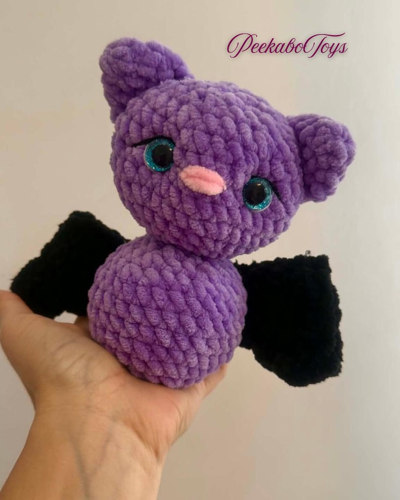 May include: A handmade purple crochet bat with black wings, sparkly blue eyes, and a pink nose. The plush toy is soft and cuddly, perfect for a child's bedroom or as a Halloween decoration. The text "Peekabo Toys" is in the upper right corner.