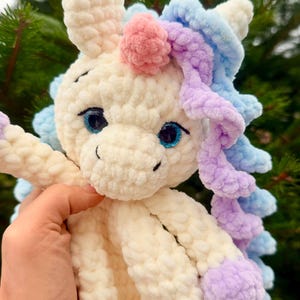 May include: A handmade, plush unicorn toy with a cream-colored body, blue eyes, and a pink horn. The unicorn has blue and purple yarn hair and accents. The toy is held in a person's hand against a blurred green background.