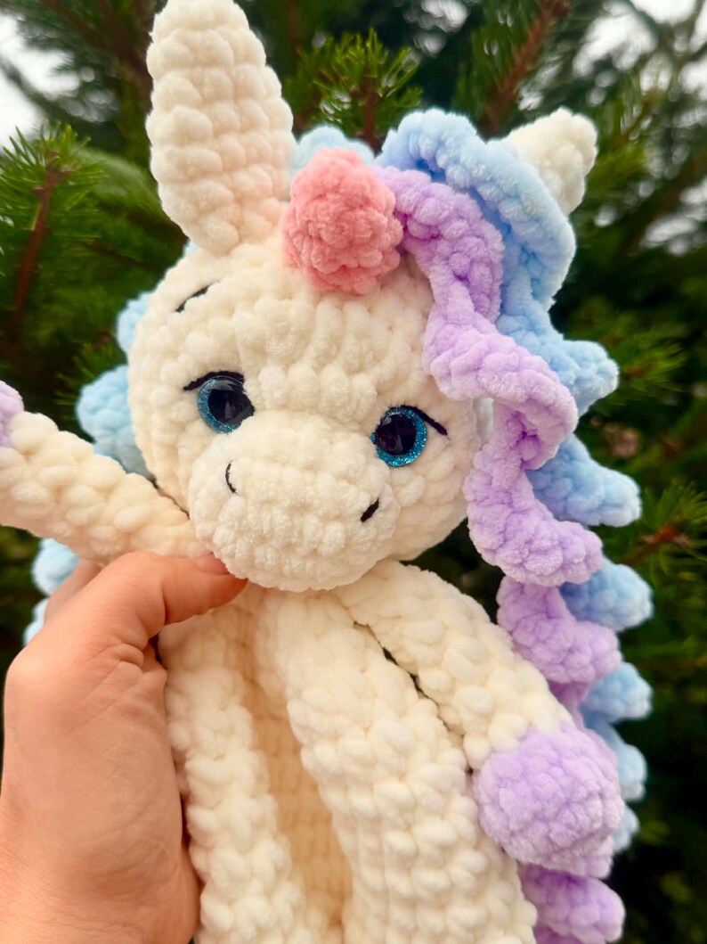 May include: A handmade, plush unicorn toy with a cream-colored body, blue eyes, and a pink horn. The unicorn has blue and purple yarn hair and accents. The toy is held in a person's hand against a blurred green background.