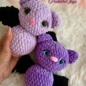 May include: Two handmade crochet bat toys in shades of purple. The bats have black wings, pink noses, and large, glittery eyes. The toys are soft and plush, perfect for children.