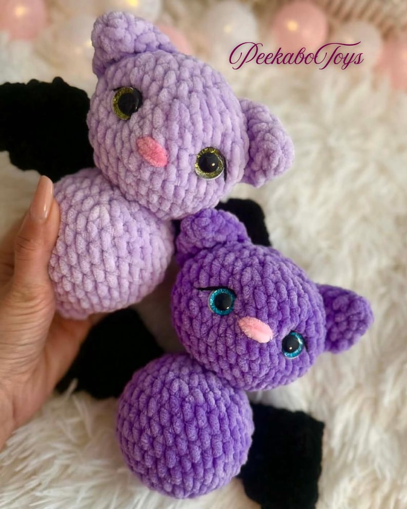 May include: Two handmade crochet bat toys in shades of purple. The bats have black wings, pink noses, and large, glittery eyes. The toys are soft and plush, perfect for children.