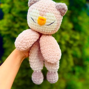 May include: A handmade crochet owl plush toy in soft pink and gray hues. The owl features closed eyes, a yellow beak, and small ear tufts. The plush toy is held against a blurred green background.