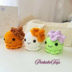 May include: Three crocheted octopus toys in orange, white, and green. Each has a ruffled headpiece in brown, orange, and lavender, respectively, and smiling faces. The toys are on a white surface, with the text "PeekaboToys" in the bottom right.