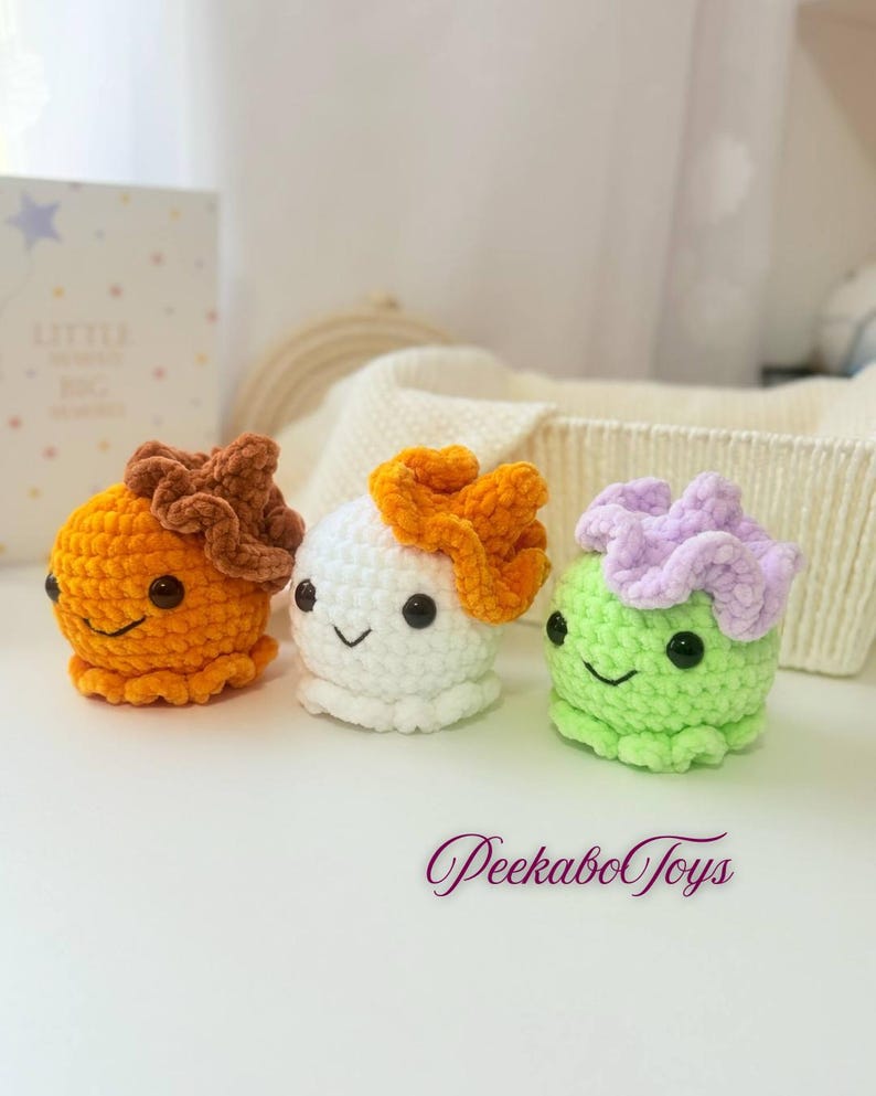 May include: Three crocheted octopus toys in orange, white, and green. Each has a ruffled headpiece in brown, orange, and lavender, respectively, and smiling faces. The toys are on a white surface, with the text "PeekaboToys" in the bottom right.