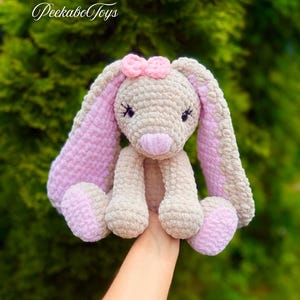 May include: A handmade crochet bunny plush toy in beige and pink. The bunny has long, floppy ears with pink lining, a pink bow, and a pink nose. The text "PeekaboToys" is in the upper left corner.