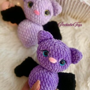 May include: Two plush, purple bat toys with black wings. The bats have large, round eyes with blue and green accents, and pink noses. The toys are handmade and have a soft, textured appearance. The text "PeekaboToys" is visible in the background.