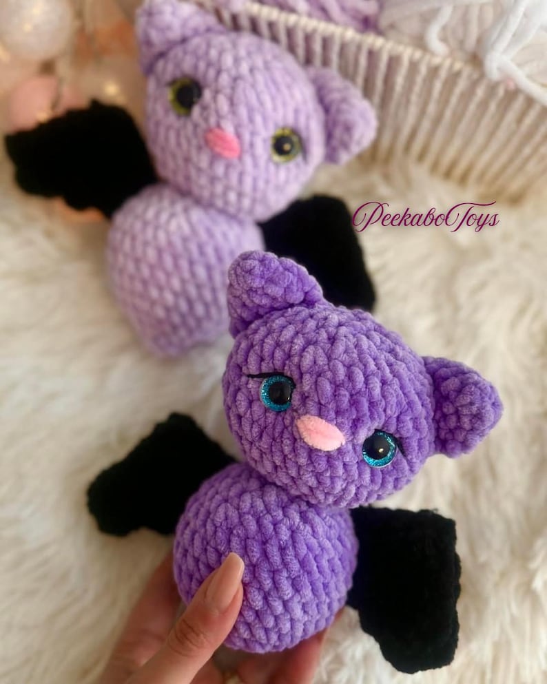 May include: Two plush, purple bat toys with black wings. The bats have large, round eyes with blue and green accents, and pink noses. The toys are handmade and have a soft, textured appearance. The text "PeekaboToys" is visible in the background.