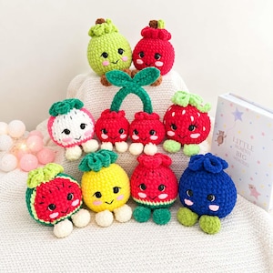Könnte beinhalten: Eine Sammlung von zehn kleinen, runden, gehäkelten Plüschfiguren in Form verschiedener Früchte, darunter Wassermelone, Ananas, Erdbeere, Blaubeere und Kirschen. Jedes Plüschtier hat gestickte Gesichtsausdrücke, rosa Wangen und kleine weiße Füße.