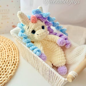 May include: A handmade, plush unicorn toy with blue, purple, and pink accents, nestled in a woven basket. The unicorn has a cream-colored body, blue eyes, and a fluffy mane. The text "PeekaboToys" is visible in the upper right corner.