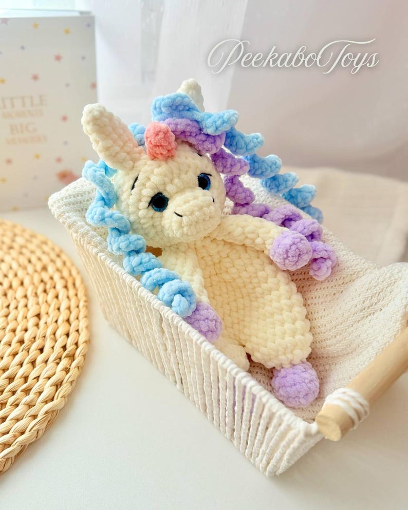 May include: A handmade, plush unicorn toy with blue, purple, and pink accents, nestled in a woven basket. The unicorn has a cream-colored body, blue eyes, and a fluffy mane. The text "PeekaboToys" is visible in the upper right corner.