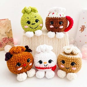May include: Five round, plush, crocheted stuffed toys with embroidered faces, rosy cheeks, and white feet. The collection includes a green sphere, a brown mug with a red handle, a dark brown toy, a white snowman-like figure, and a tan textured sphere.