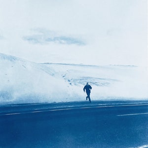 May include: A person in a dark jacket runs across a paved road in front of a snow-covered landscape.
