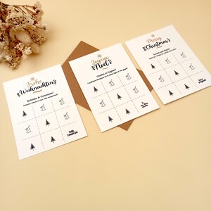 May include: Three Christmas scratch-off cards with gold and black text. The cards feature a grid with Christmas tree and bell illustrations. The text includes "Merry Christmas", "Joyeux No&euml;l", and "Frohe Weihnachten". A brown envelope is behind the cards.