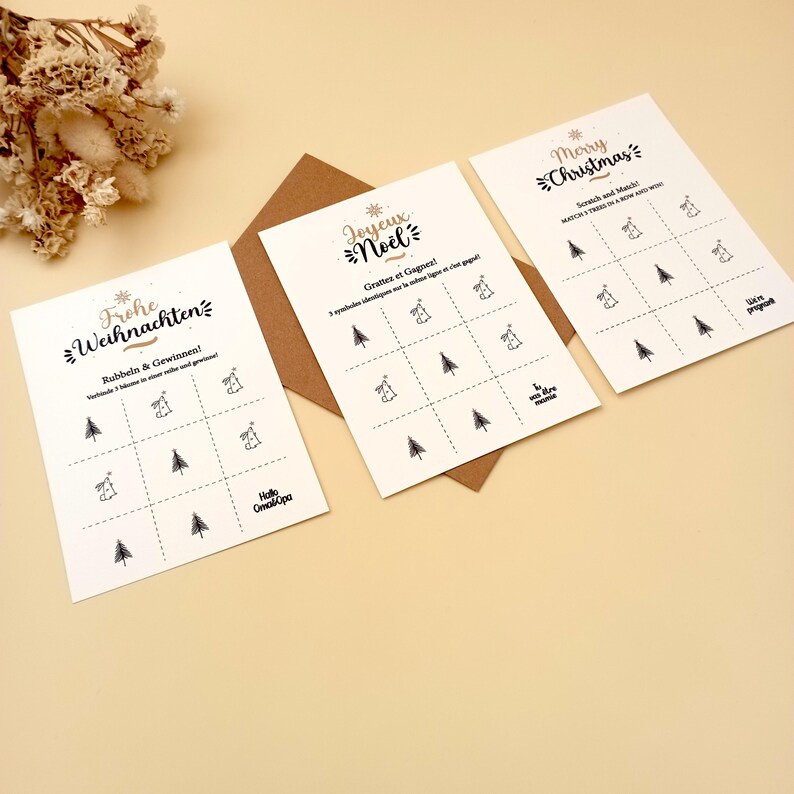 May include: Three Christmas scratch-off cards with gold and black text. The cards feature a grid with Christmas tree and bell illustrations. The text includes "Merry Christmas", "Joyeux No&euml;l", and "Frohe Weihnachten". A brown envelope is behind the cards.