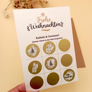 May include: A cream-coloured Christmas scratch-off card with gold foil circles and the German text "Frohe Weihnachten." The card features a tic-tac-toe style layout with Christmas tree illustrations. The text "Rubbeln & Gewinnen!" is also visible.