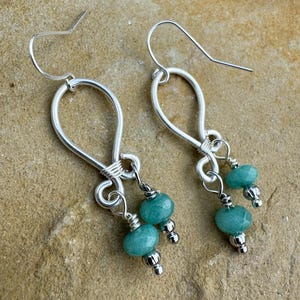 May include: A pair of handcrafted silver earrings. Each earring features a looped design with a wire-wrapped accent, suspending two turquoise-colored gemstone beads and silver bead accents. The earrings have a hook closure.