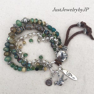 May include: A multi-strand bracelet featuring green, brown, and blue beads, accented with silver-toned metal beads and chain. The bracelet includes a brown leather cord and decorative charms, including a bird and a leaf. The text "JustJewelrybyJP" is visible.