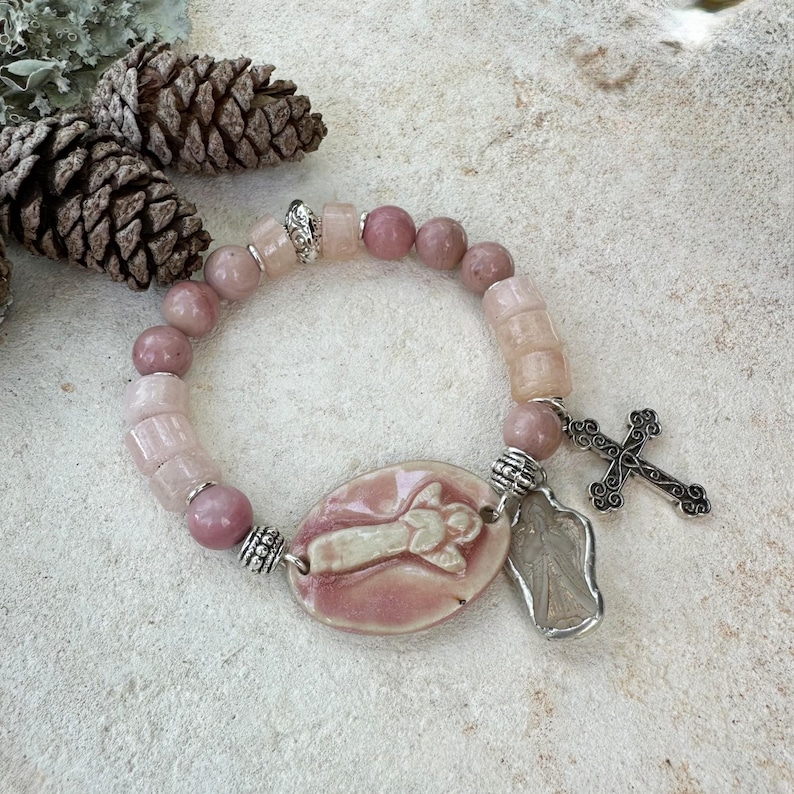 May include: A pink beaded bracelet with a ceramic oval pendant featuring a cross design. The bracelet includes a silver cross charm and a silver charm of a saint. The beads are various shades of pink.