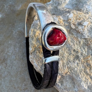 May include: Silver-tone bracelet with a red stone centerpiece and a black leather band. The bracelet features a textured leather band and a polished silver clasp. The red stone is set in a silver bezel.