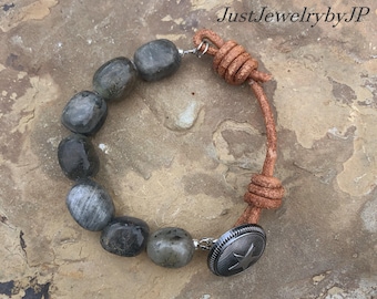 May include: A bracelet featuring gray stone beads and a brown leather cord. The bracelet has a silver-tone button clasp with a star design. The text "JustJewelrybyJP" is visible in the image.
