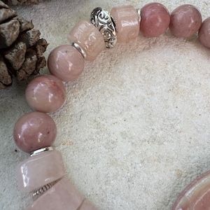 May include: A close-up of a bracelet featuring pink and silver beads. The bracelet has a mix of round and rectangular beads in varying shades of pink. Silver spacers with intricate designs separate some of the beads. The bracelet is on a light-colored surface.