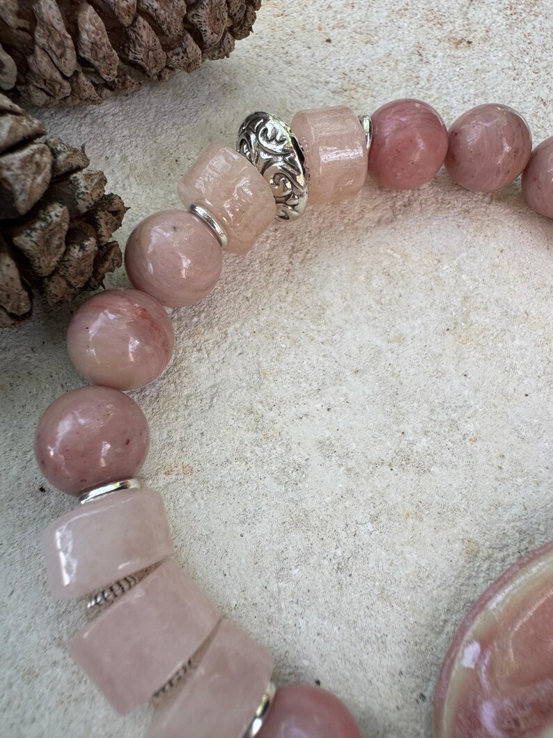 May include: A close-up of a bracelet featuring pink and silver beads. The bracelet has a mix of round and rectangular beads in varying shades of pink. Silver spacers with intricate designs separate some of the beads. The bracelet is on a light-colored surface.