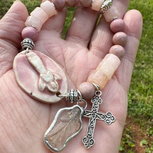 May include: A pink beaded bracelet with a religious theme. The bracelet features a variety of pink beads, including round and rectangular shapes. It has a silver cross charm and an angel charm. The bracelet is held in a hand.