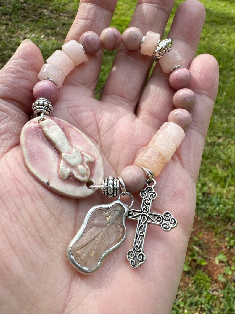 May include: A pink beaded bracelet with a religious theme. The bracelet features a variety of pink beads, including round and rectangular shapes. It has a silver cross charm and an angel charm. The bracelet is held in a hand.