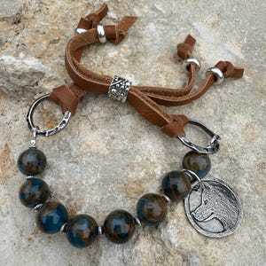 May include: A handcrafted bracelet featuring large, round beads in shades of blue and brown, strung together with silver-toned spacers. The bracelet includes a brown leather strap with silver accents and a circular pendant with a wolf's head.