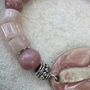 May include: A close-up of a pink beaded necklace. The necklace features a variety of pink beads, including round and rectangular shapes, and a large, oval-shaped pendant. Silver-colored metal accents are visible between the beads.
