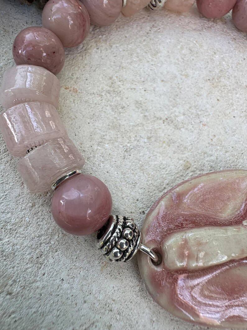 May include: A close-up of a pink beaded necklace. The necklace features a variety of pink beads, including round and rectangular shapes, and a large, oval-shaped pendant. Silver-colored metal accents are visible between the beads.