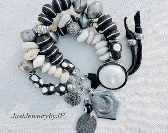 May include: A multi-strand bracelet featuring black and white beads, silver accents, and charms. The charms include a moon, a sun face, and an oval pendant. The bracelet is made with a black leather cord.