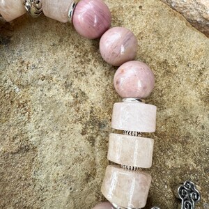 May include: Close-up of a beaded bracelet featuring pink and light pink gemstones. The bracelet includes round and rectangular beads, separated by silver-colored metal accents. A decorative silver cross charm is visible.