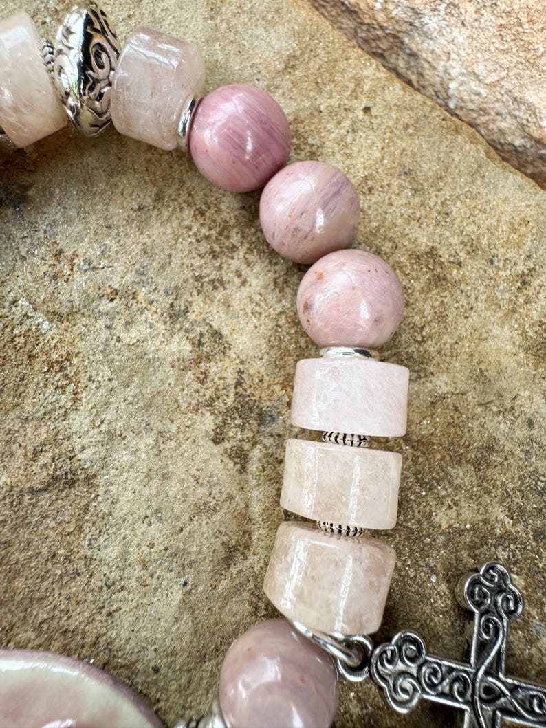 May include: Close-up of a beaded bracelet featuring pink and light pink gemstones. The bracelet includes round and rectangular beads, separated by silver-colored metal accents. A decorative silver cross charm is visible.