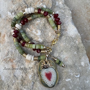 May include: A layered necklace with green, white, and red beads, accented with gold-colored metal. The pendant is a heart shape with a red center and a gold-colored crown. The text "JustJewelrybyJP" is visible.