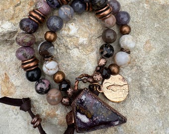 May include: A handcrafted bracelet featuring a mix of gemstone beads in shades of purple, gray, and brown, accented with copper-colored spacers. A triangular pendant and a round charm with a design hang from the bracelet, along with a brown leather cord.