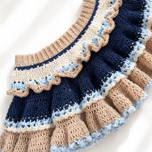 May include: A handmade crochet skirt featuring a tiered design with alternating colors. The skirt showcases a combination of navy blue, beige, and light blue, with a scalloped edge. The craftwork is visible in the close-up shot.