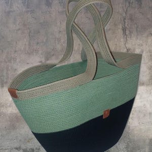May include: A large, green and black woven tote bag with brown leather handles. The bag is made of thick rope and has a wide, open top.