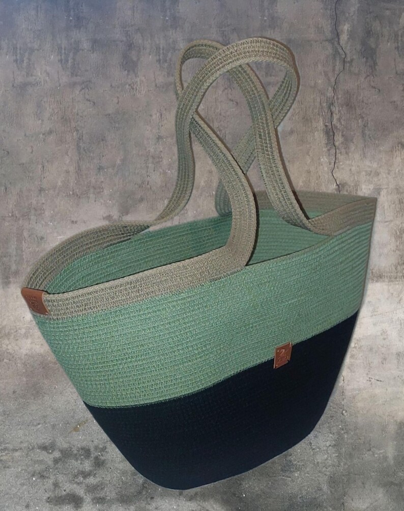 May include: A large, green and black woven tote bag with brown leather handles. The bag is made of thick rope and has a wide, open top.