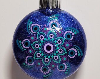 May include: A round, blue glitter Christmas ornament with a detailed mandala design in teal, purple, and green. The ornament has a silver metal cap and a black string for hanging. The mandala features concentric circles and dot patterns.