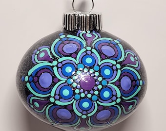 May include: A round, clear glass Christmas ornament with a black glitter background. The ornament features a symmetrical mandala design in shades of purple, teal, and blue. The ornament is suspended by a black cord and silver metal cap.