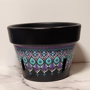 May include: A black plant pot featuring a hand-painted dot art pattern in shades of purple, teal, and white. The design consists of symmetrical teardrop and circular motifs arranged in rows around the exterior. The pot includes vertical drainage slits.