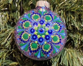 May include: Glittery Christmas ornament with a hand-painted mandala design. The ornament features a pattern of blue, green, and yellow dots and floral shapes. The ornament is round and has a silver cap.