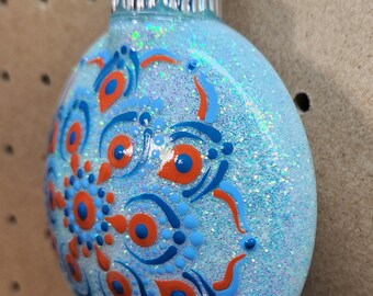 May include: A light blue, glitter-covered Christmas ornament with a silver cap. The ornament features a detailed design of blue and orange dots and curved lines, creating a symmetrical pattern. The ornament is hanging from a silver hook.