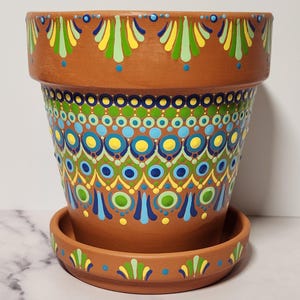 Puede incluir: Una maceta de color terracota con un platillo a juego. La maceta está adornada con un diseño pintado a mano con patrones circulares y de gotas azules, amarillos y verdes. El borde y el platillo tienen un motivo repetitivo en forma de hoja.