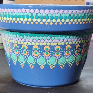 Puede incluir: Una maceta de terracota azul con un diseño de mandala pintado a mano. El diseño presenta un degradado de puntos verdes, amarillos y morados, con un borde de puntos naranjas y verdes. La maceta tiene un agujero de drenaje.